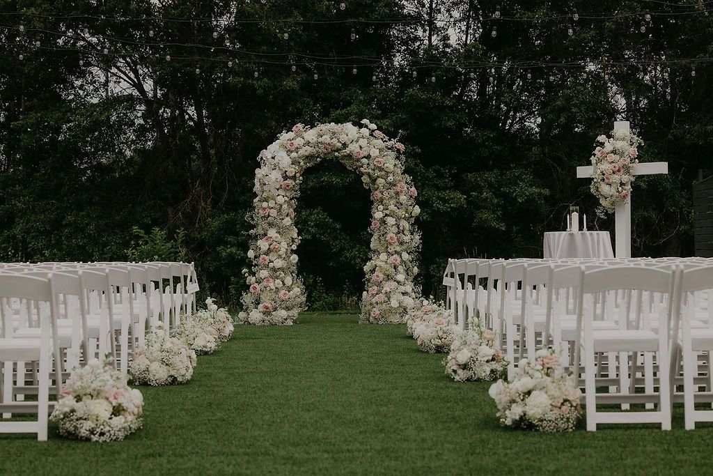 Outdoor wedding ceremony with white floral arch and chairs on lawn